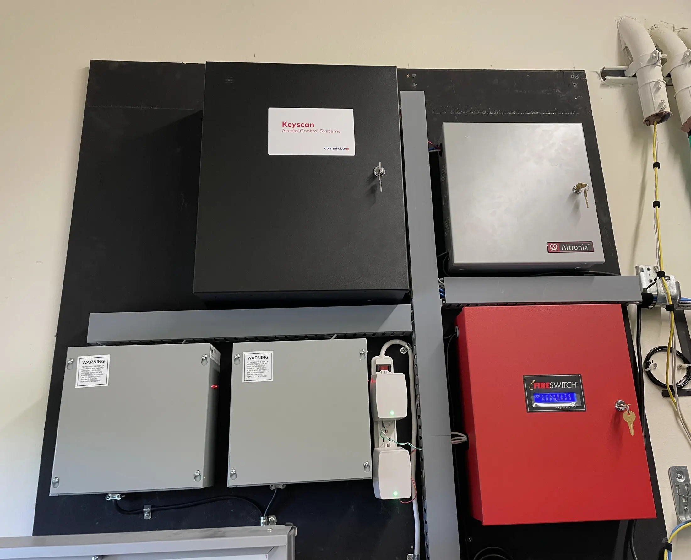 A security backboard carrying a locked black access control enclosure, three grey power supply cabinets and a red power controller that releases the door locks on a fire alarm signal, its eight channel display lit, with green, white and yellow cable bundles strapped in along the edge of the board.
