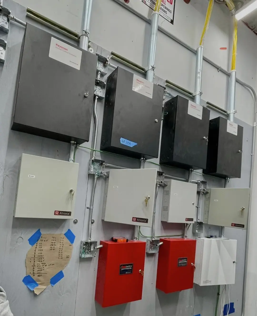 Four access control enclosures in a row above four power supply cabinets and two red modules that release the door locks on an alarm signal, all fed by rigid conduit with green bonding jumpers, and a handwritten door schedule taped to the wall beside them.