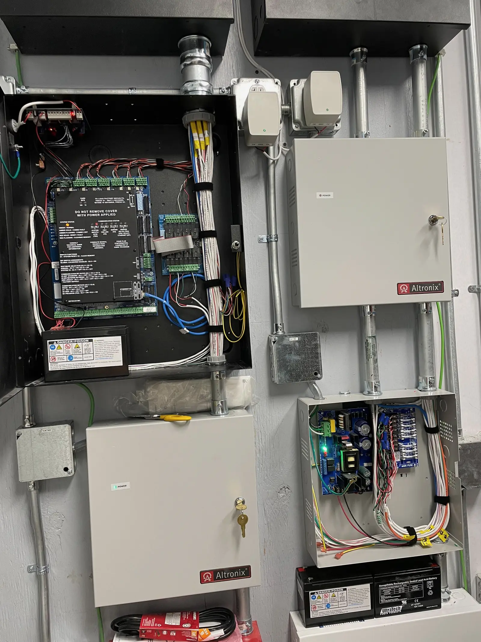 An access control head end part way through installation, the controller enclosure open to show the main board wired to labelled terminal strips, a reader interface module on ribbon cable and a sealed battery below, alongside two closed power supply cabinets and a third opened onto its board and a formed loop of colour coded conductors, all fed by EMT conduit through junction boxes labelled with the circuit number.