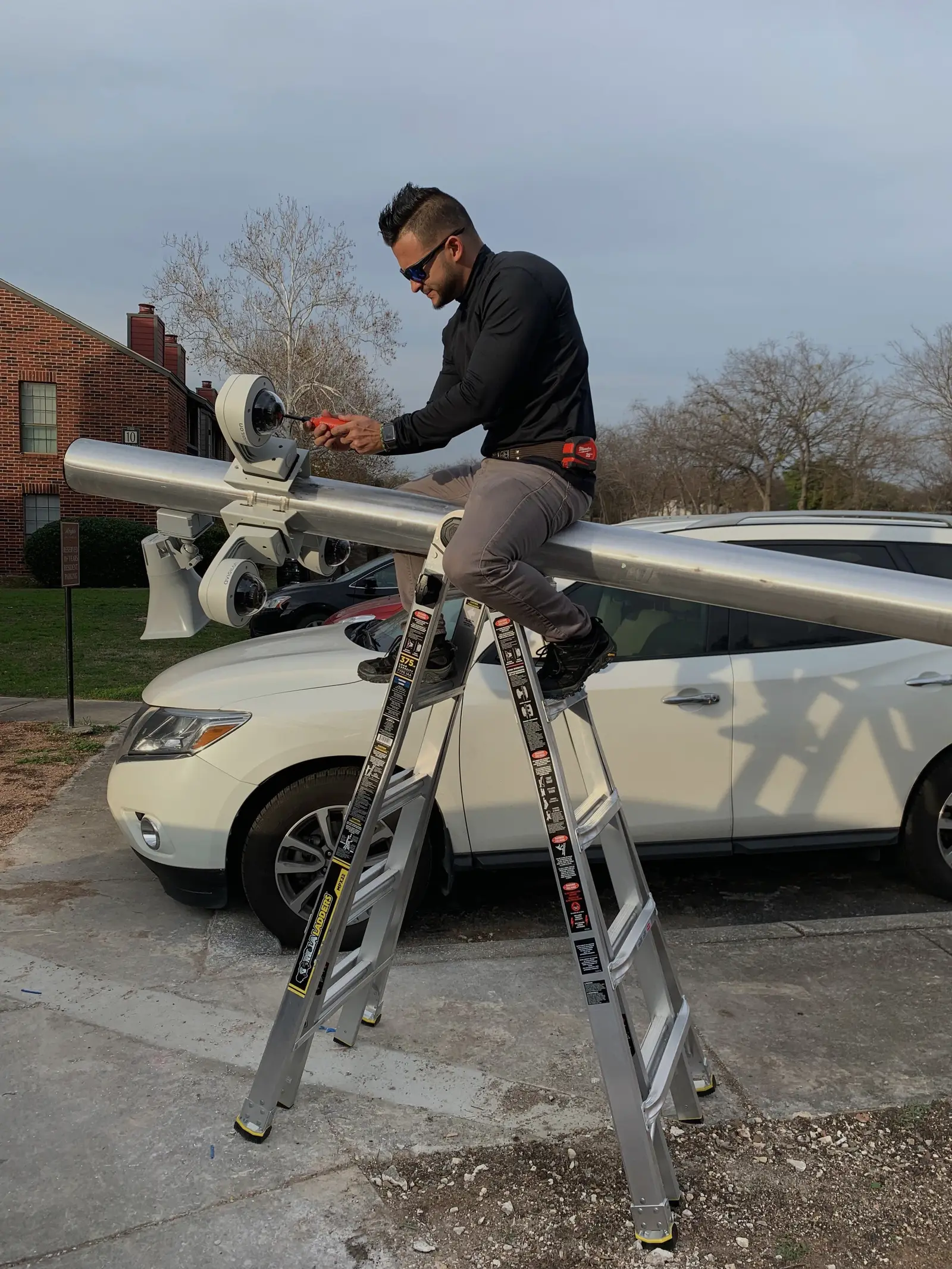 A technician sitting on a ladder to aim a four camera array mounted on a steel pole above a parking area.