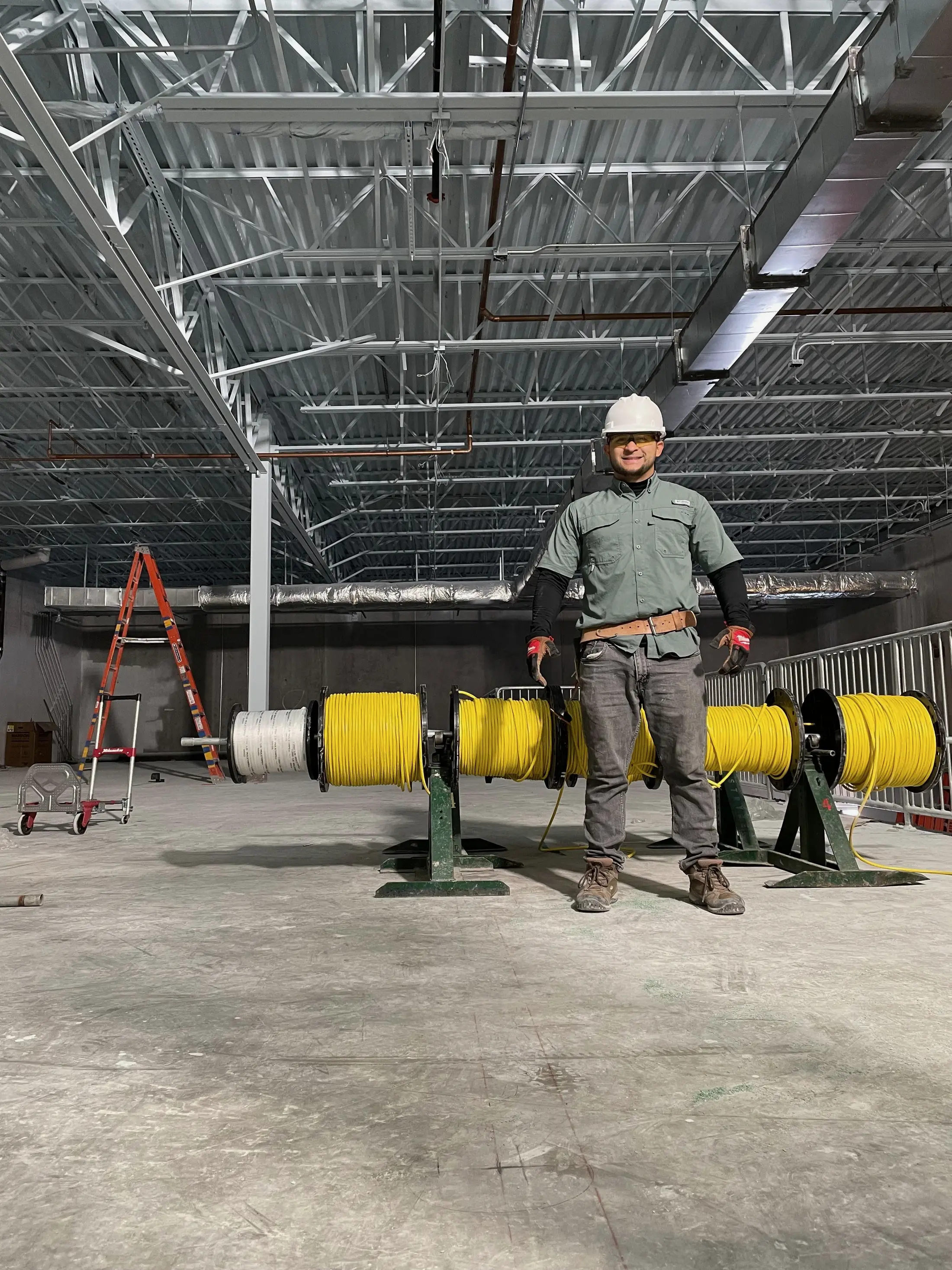 A technician in a hard hat standing in front of four cable reels on jack stands inside a building under construction, with exposed steel joists and ductwork overhead.