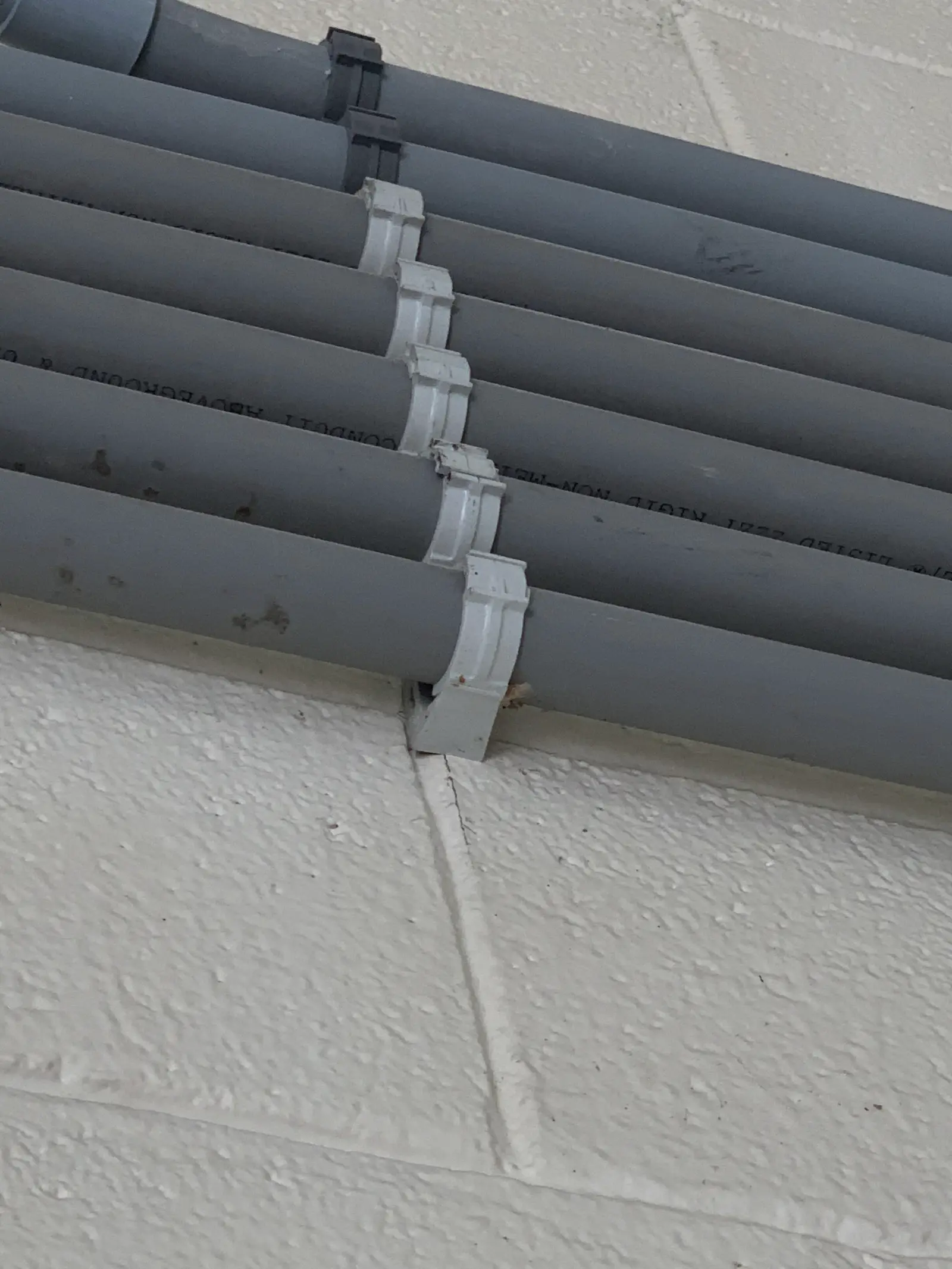 A bank of grey rigid non metallic conduits running side by side across a painted block wall, each pipe held by its own clamp with the clamps stepping down one after another across the run, and the printed conduit legend still legible along the pipes.