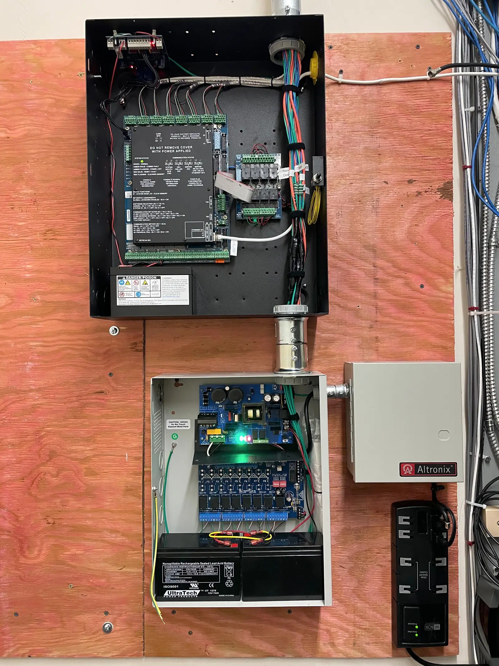 A door controller panel and a battery backed power supply mounted on a plywood backboard, wired with colour coded conductors and dressed into flexible conduit.