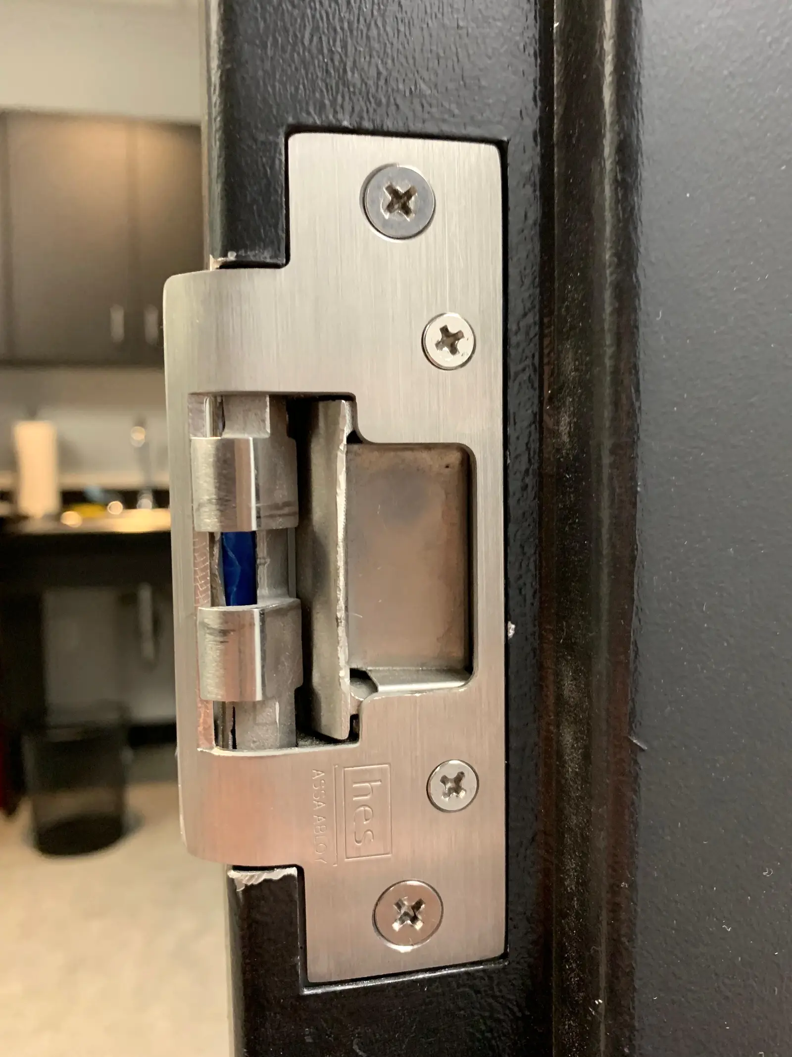 Close up of a brushed stainless electric strike set into a dark steel door frame, its four screws seated flush, the keeper and solenoid visible in the open cutout with the blue coil lead behind them, and an out of focus interior beyond the doorway.