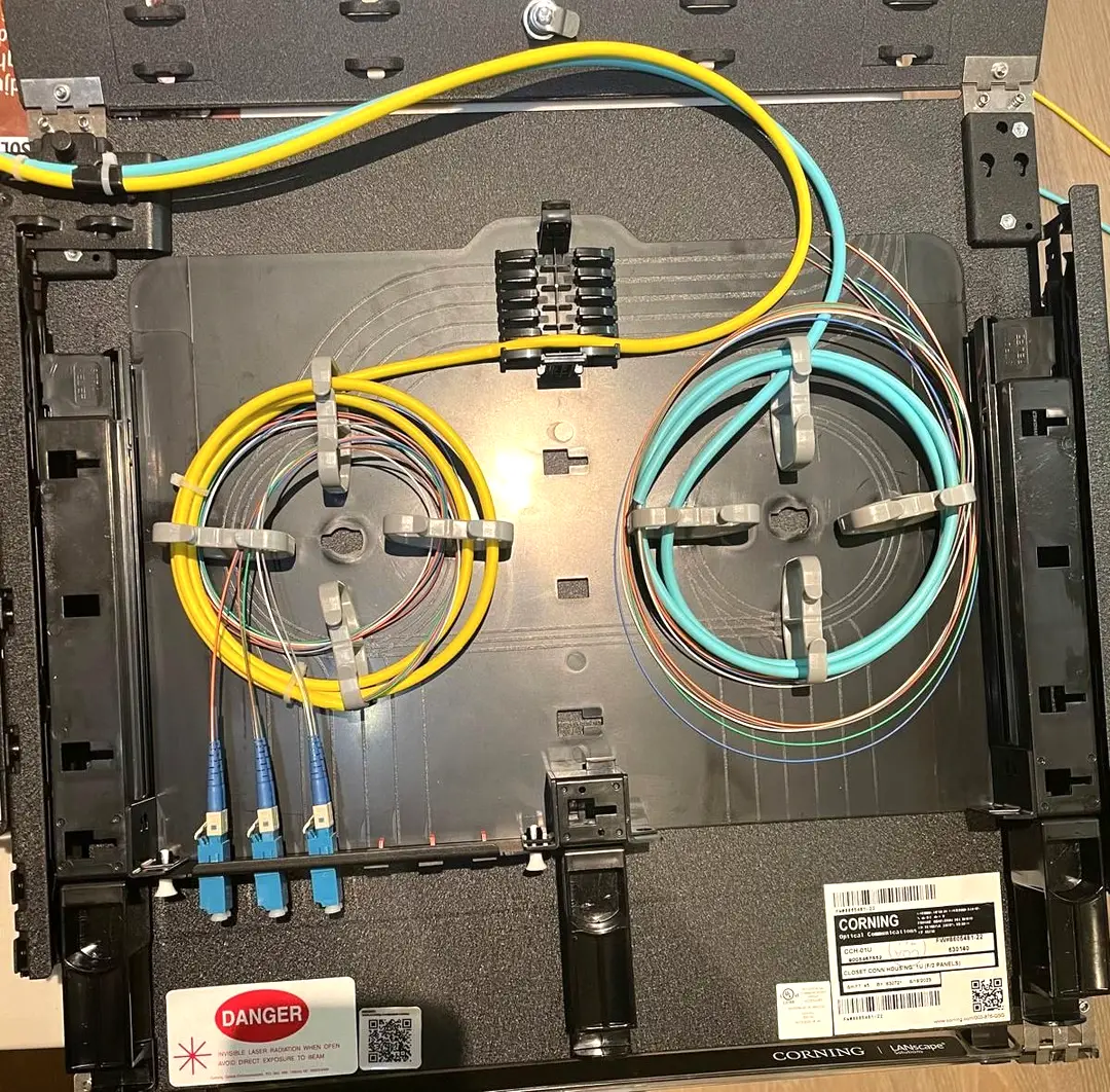 An open fiber optic housing with a yellow single mode jumper and an aqua multimode jumper coiled on the routing tray under grey retaining clips, colour coded fibers fanning out to three blue duplex LC connectors seated in the adapter panel beside a laser radiation warning label.