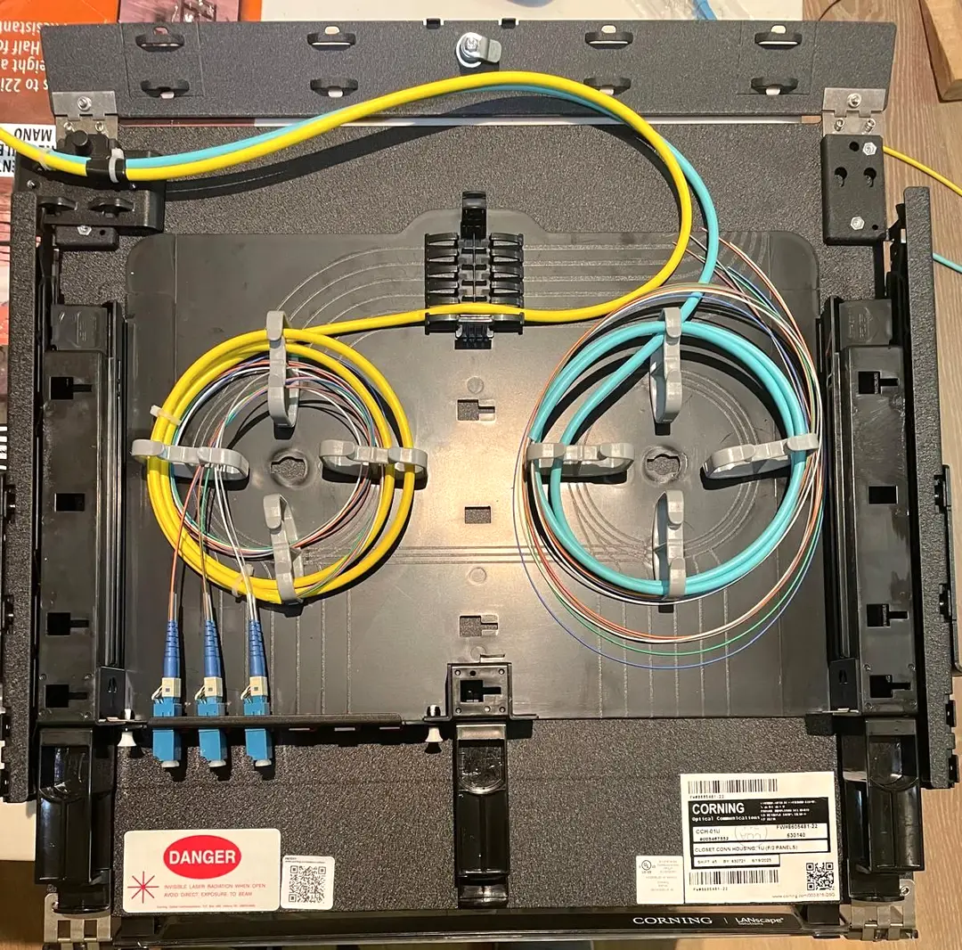 A rack mount fiber connector housing opened flat, with a yellow single mode pigtail bundle coiled on the left splice tray and an aqua multimode bundle on the right, the colour coded fibers clipped into splice holders, three blue SC connectors seated in the adapter panel and a laser radiation warning label beside them.
