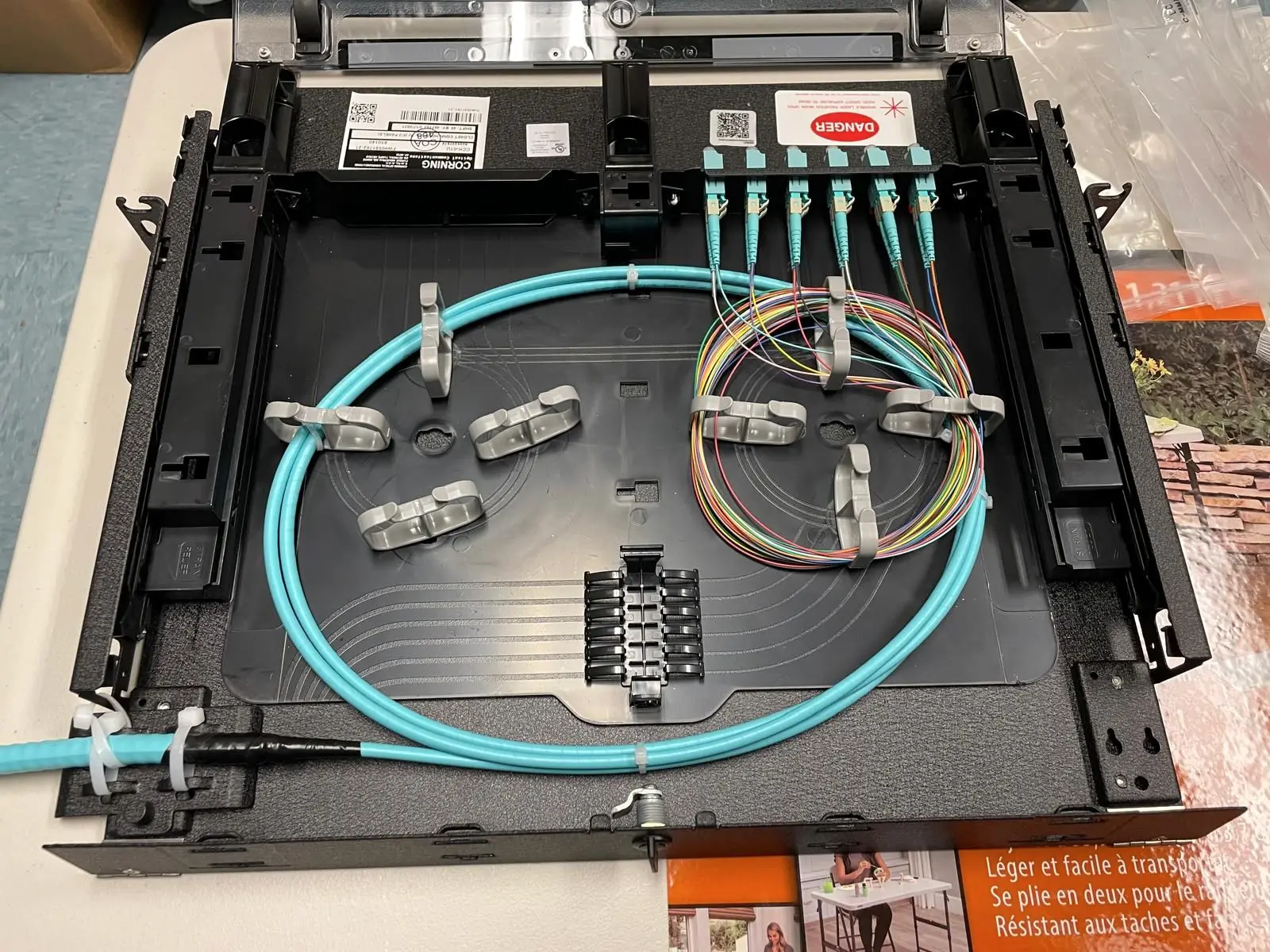 An open fiber optic wall housing on a bench, with an aqua multimode trunk coiled on the splice tray, colour coded pigtails, and six aqua LC connectors seated in the adapter panel.