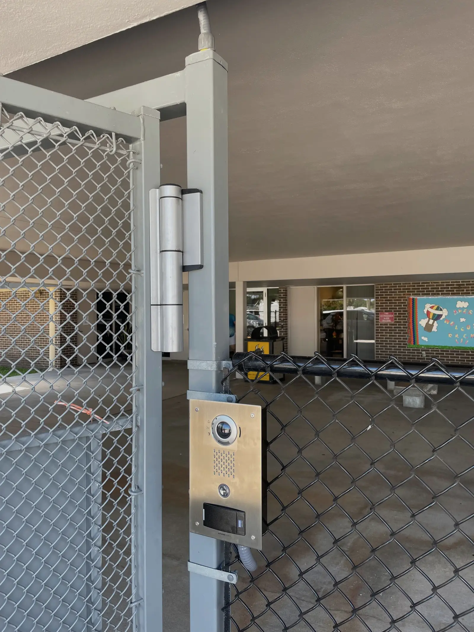 A stainless video intercom with a card reader mounted on the post of a fenced pedestrian gate, with a heavy duty gate hinge above it.