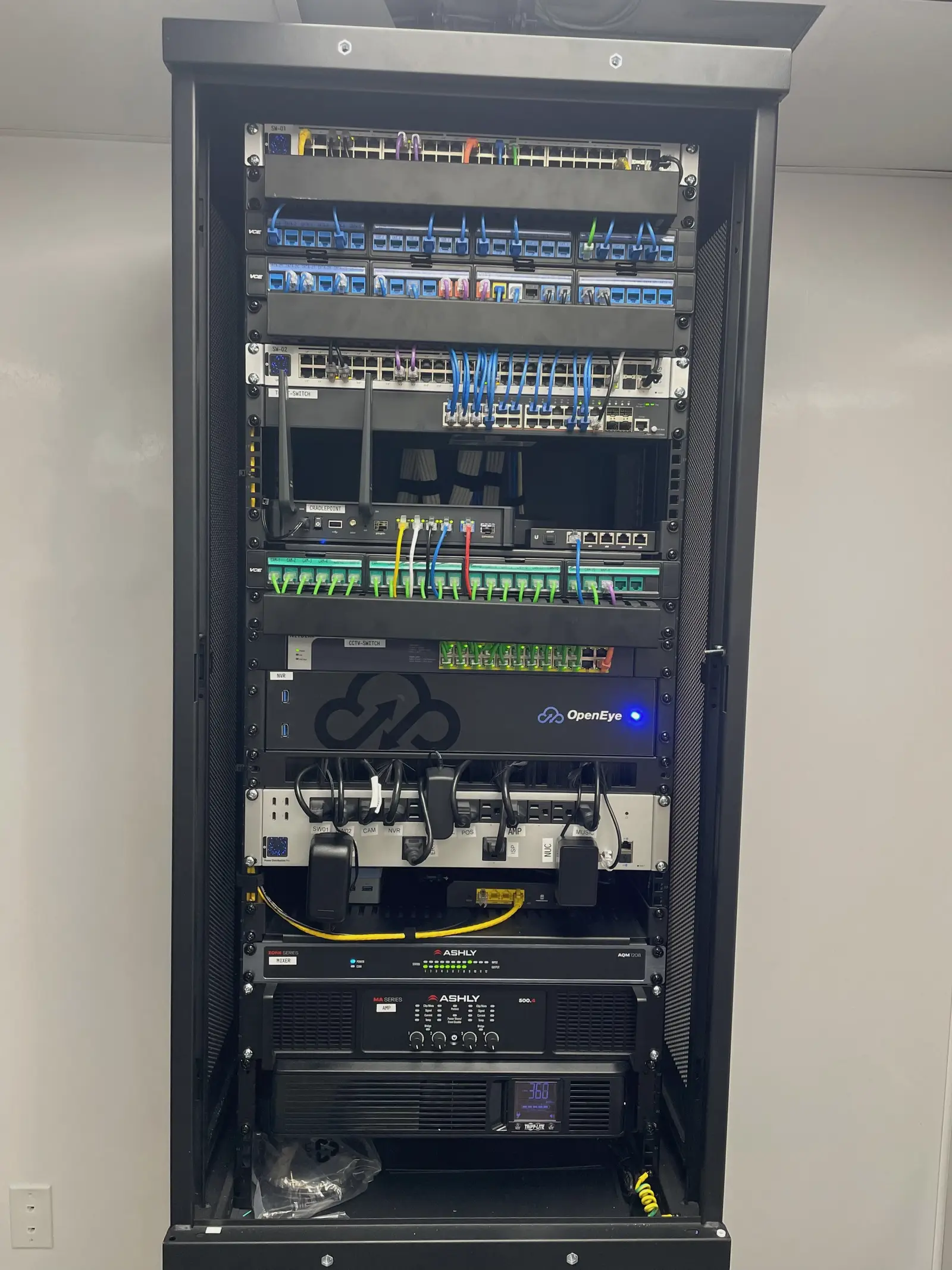 A floor standing network rack seen straight on, with two named switches, patch panels marked port by port, a camera panel wired entirely in green, a video recorder, a power strip with every outlet named, an audio mixer, an amplifier, and a battery backup at the base.