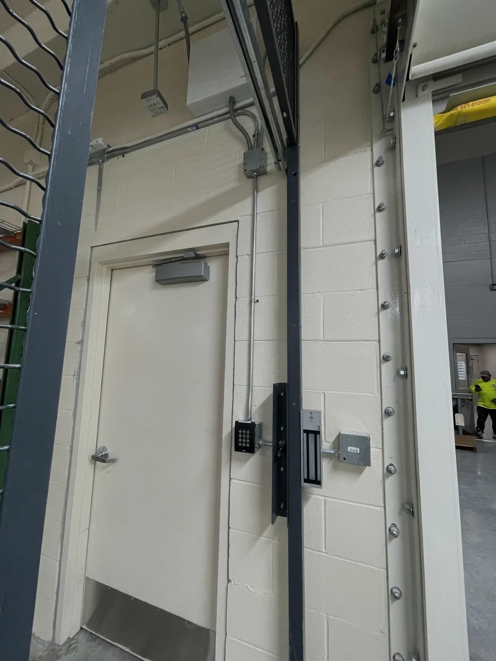A magnetic lock and keypad reader installed on the frame of a caged entrance in a warehouse, with conduit run up to a junction box.