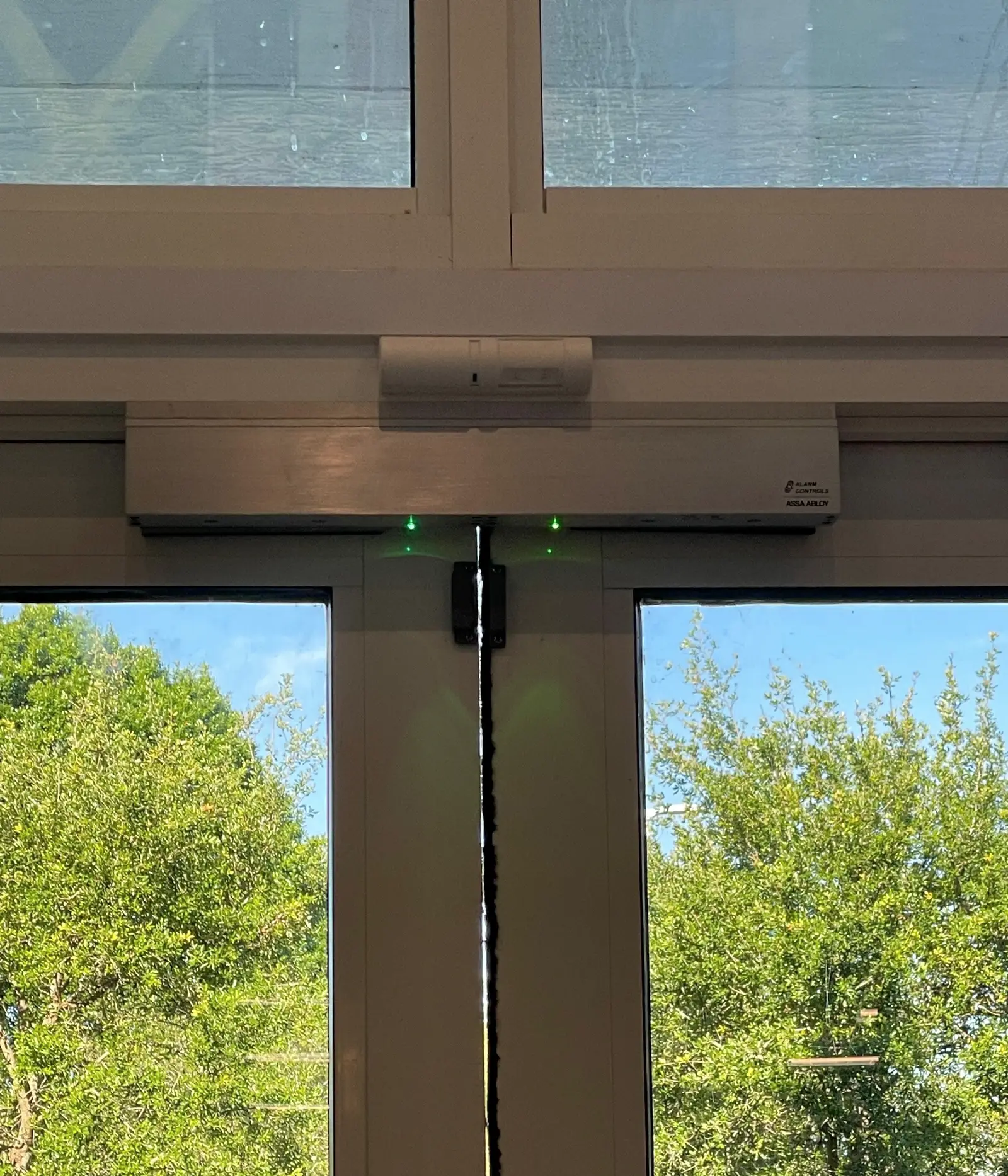 A magnetic lock bar spanning the head of a pair of aluminium framed glass doors, with two green status lights showing it energised, a request to exit sensor mounted above on the frame, and a two piece surface contact set either side of the gap where the leaves meet.
