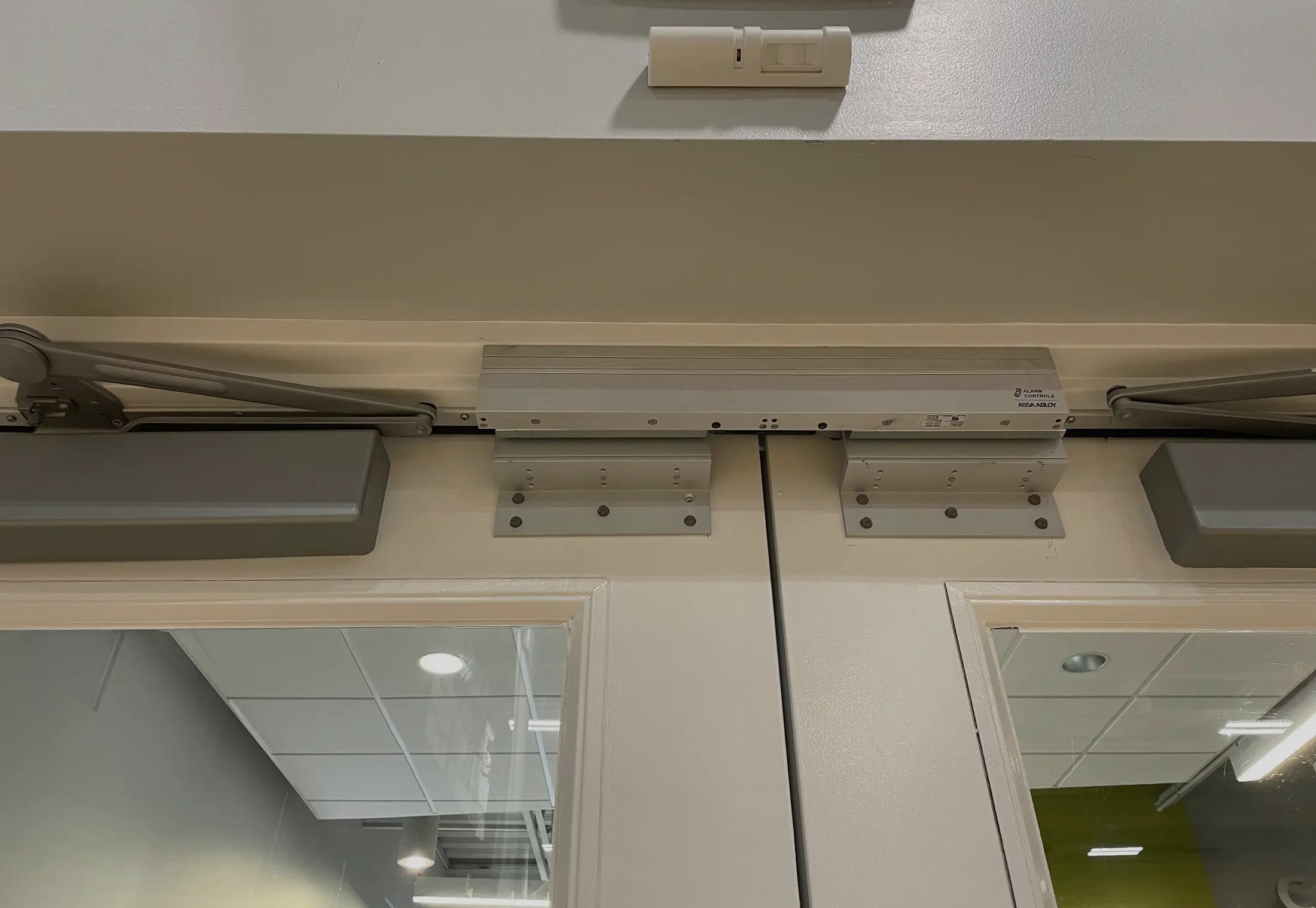 A magnetic lock mounted across the head frame of a pair of doors, spanning both leaves on two bracket plates, with a request to exit motion sensor on the wall above and an overhead closer arm on each leaf.