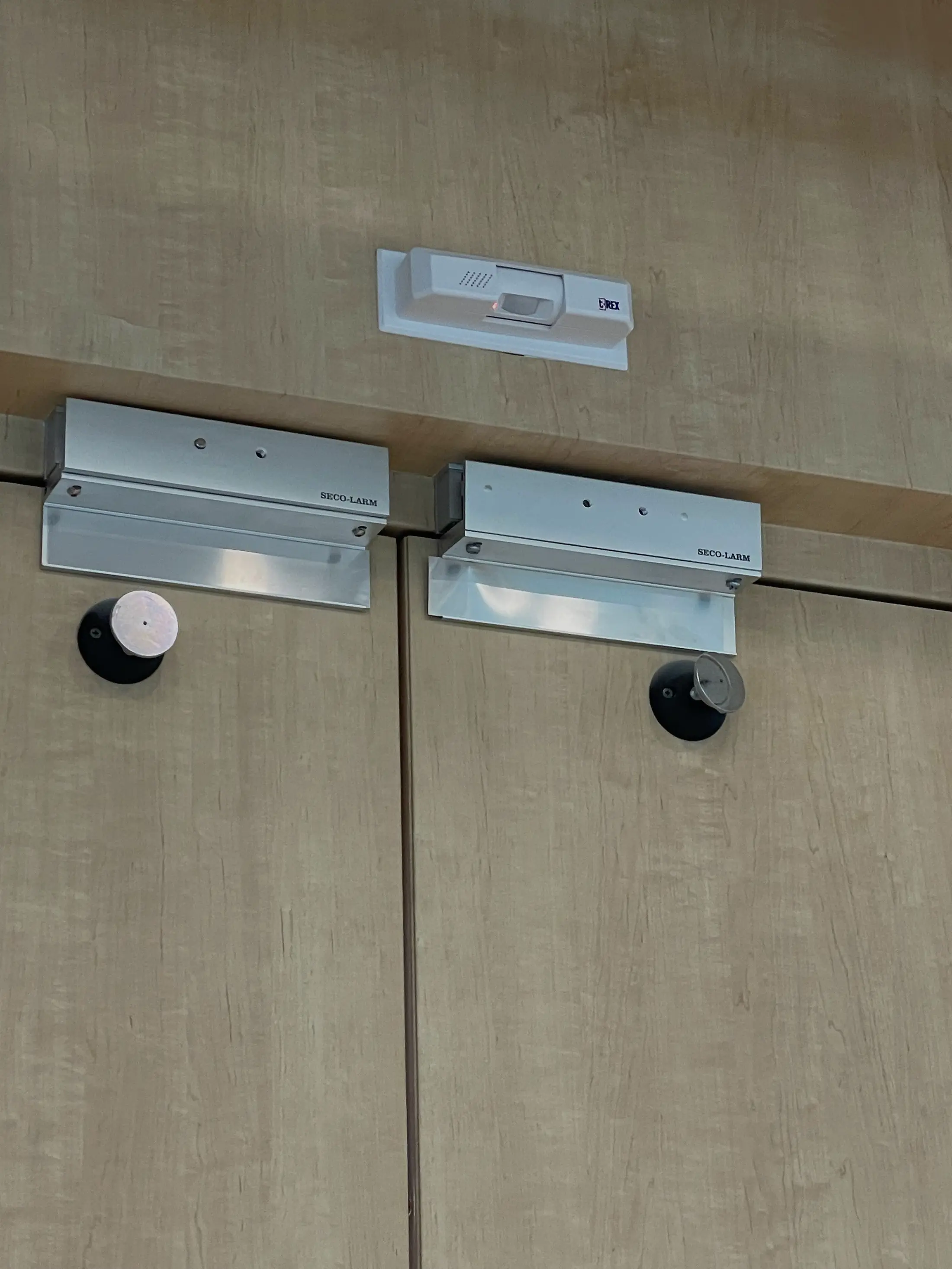 Two magnetic locks with door position sensors installed on the head frame above a pair of wooden doors.