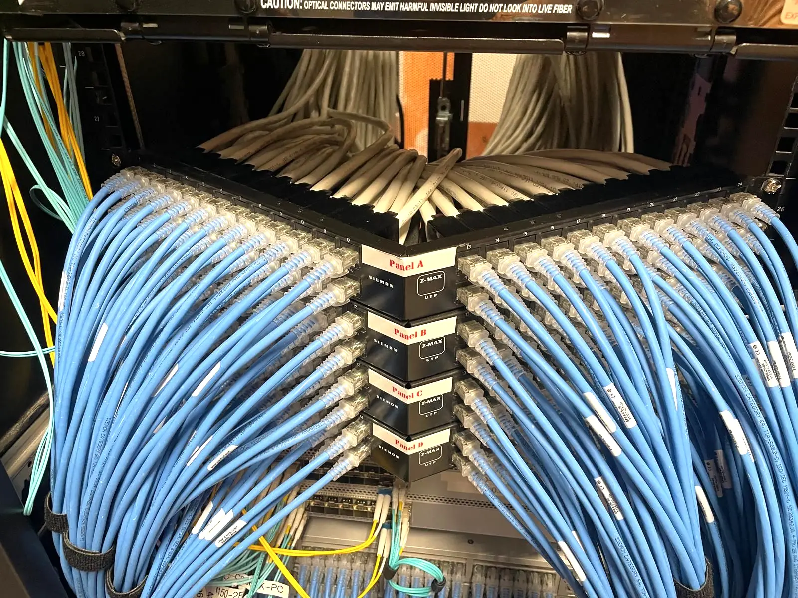 Hundreds of blue patch cords fanning out from four labelled copper patch panels inside a network rack, with a grey cable trunk above and fiber pigtails at the side.