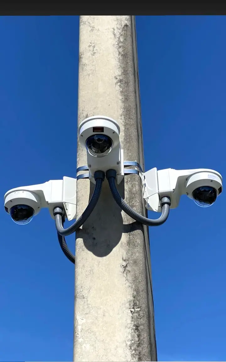 Three surveillance cameras clamped to a concrete pole and joined by flexible conduit whips, seen from below against a clear sky.