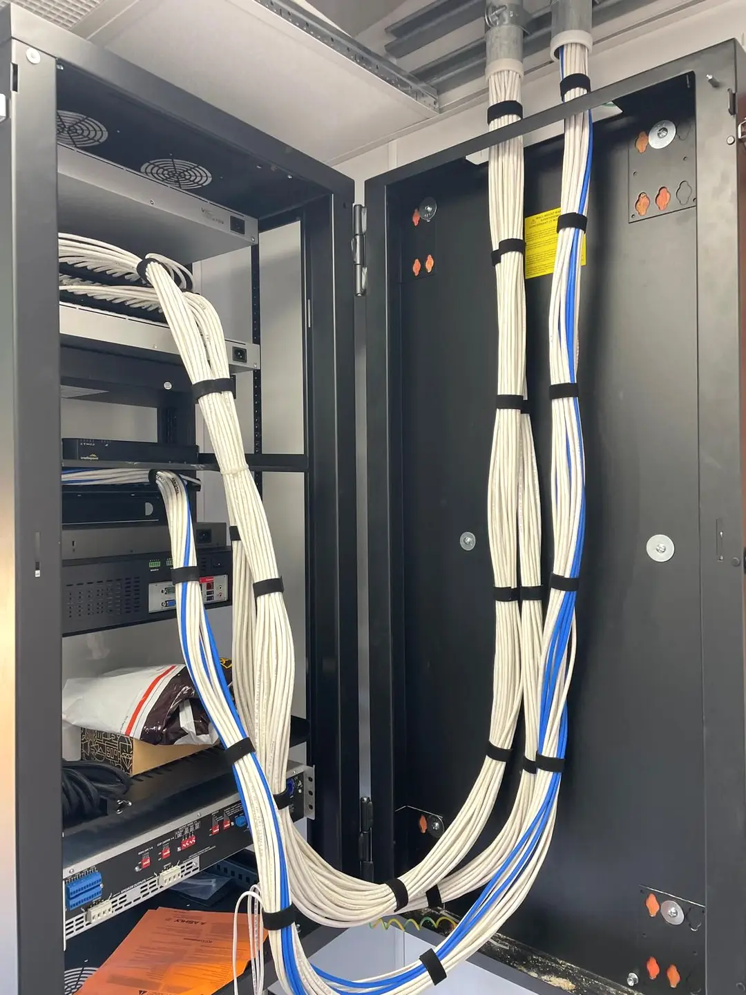 White low voltage cable and blue network cable dropping out of ceiling conduit sleeves, strapped with hook and loop at even intervals and swept through a long service loop into an open wall mount cabinet holding a cellular router and rack mounted audio equipment.