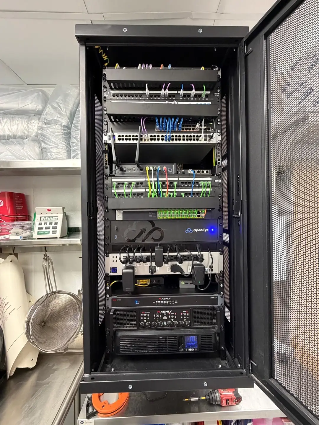 A floor standing network rack with patch panels, switches, a cellular router, a video recorder, labelled power outlets, an audio mixer, an amplifier and a UPS.
