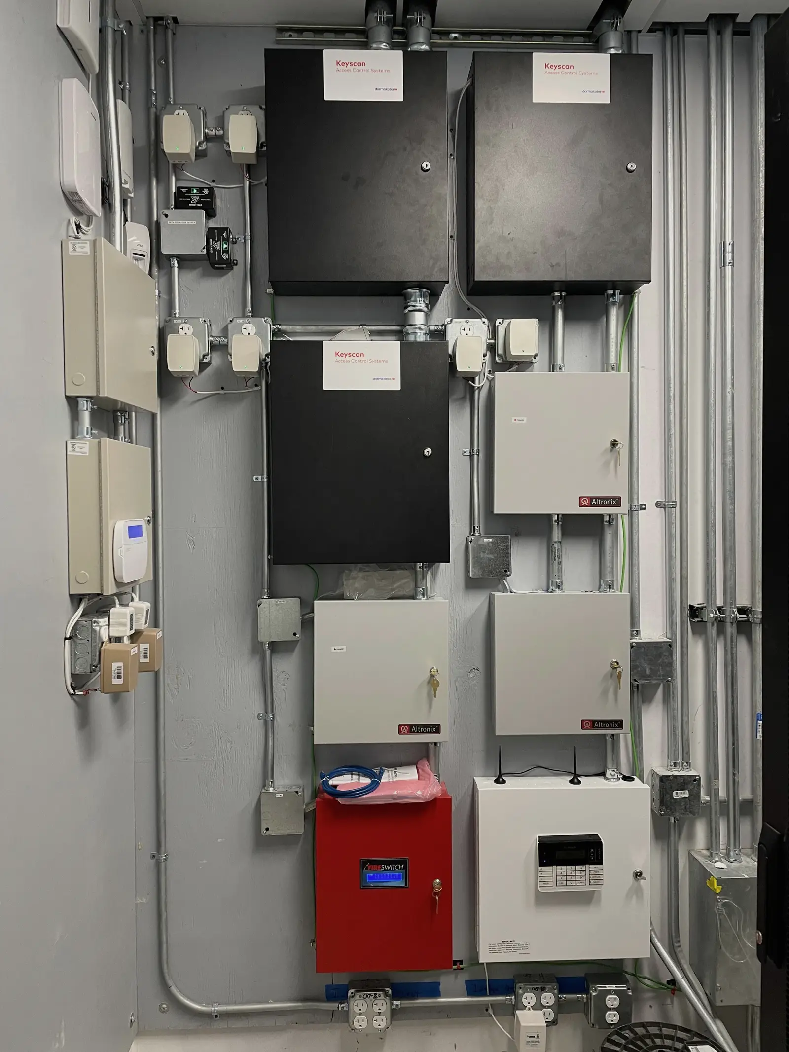 A security equipment backboard carrying three access control enclosures, four power supplies, a red power module that releases the door locks on an alarm signal, and an alarm keypad cabinet, all fed by rigid conduit.