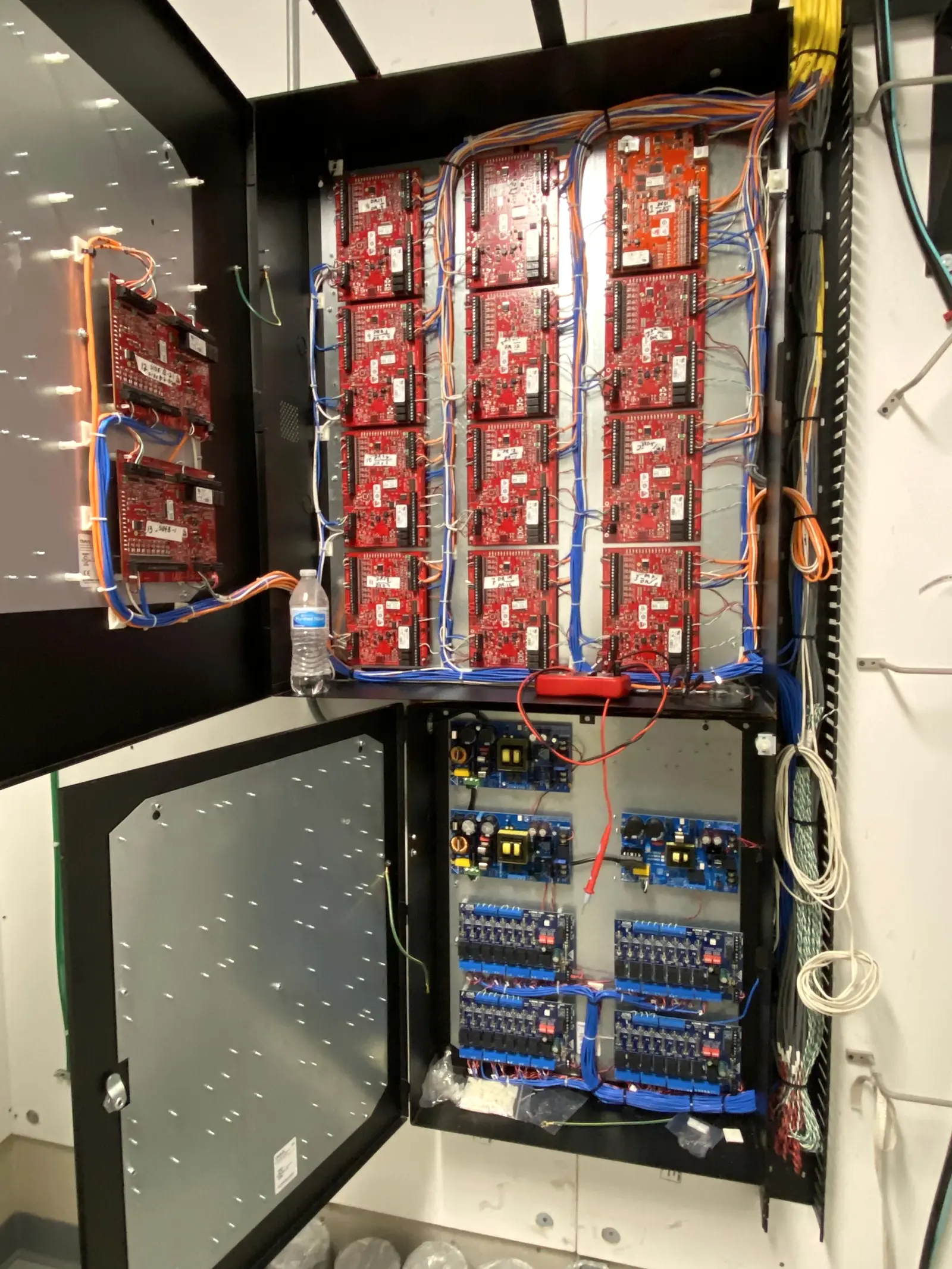 An access control enclosure opened to show twelve door controller boards wired with labelled blue and orange conductors, above a separate power distribution section.