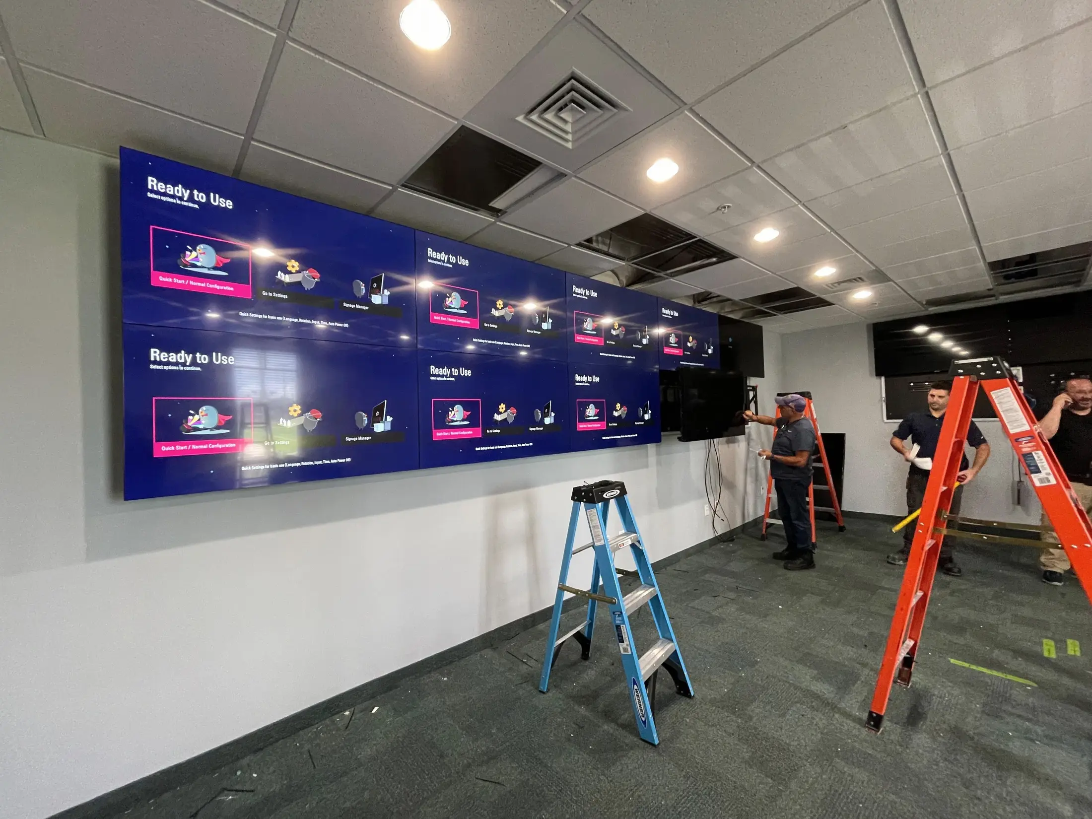 Three technicians on step ladders aligning an eight panel video wall on a dark mounting backer in an empty office space.
