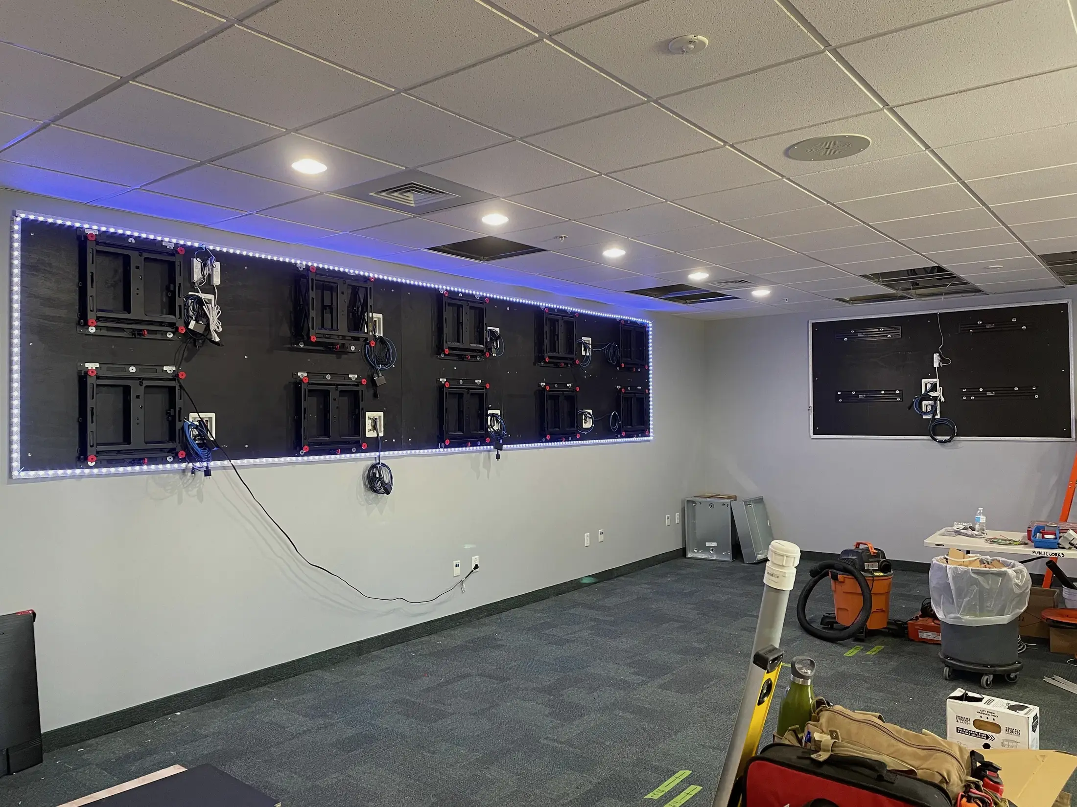 A video wall backer panel fitted with ten display brackets and LED edge lighting, with cabling coiled ready at each mount, in a room still being fitted out.